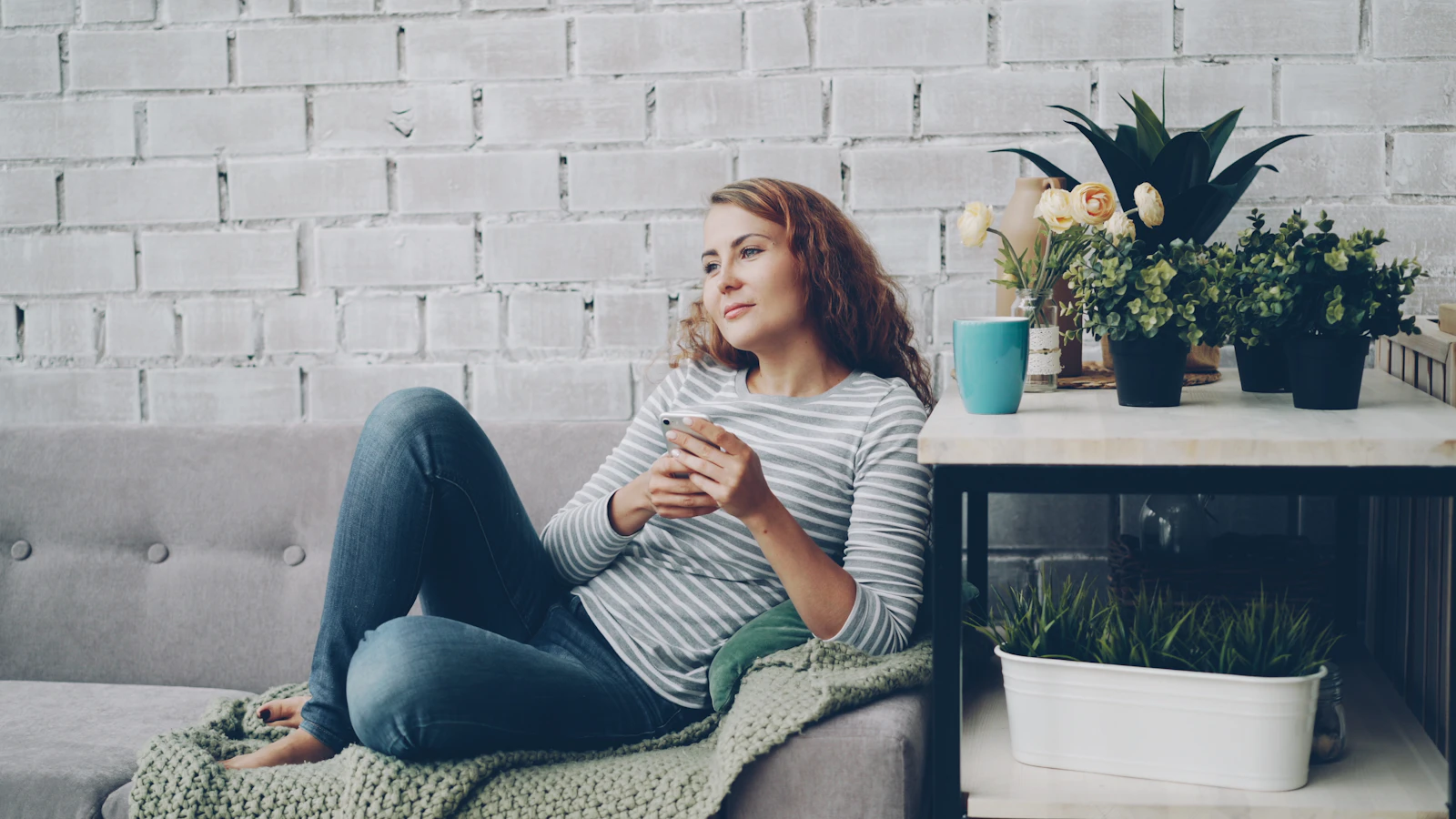 Vrouw zit met een kop thee op de bank bij een lichte muur met planten.