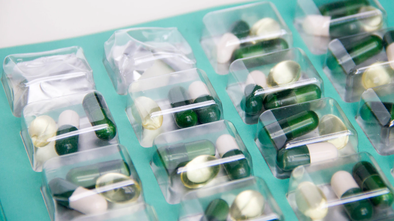 Groen-witte capsules in doorzichtige doordrukverpakking op een turquoise ondergrond.