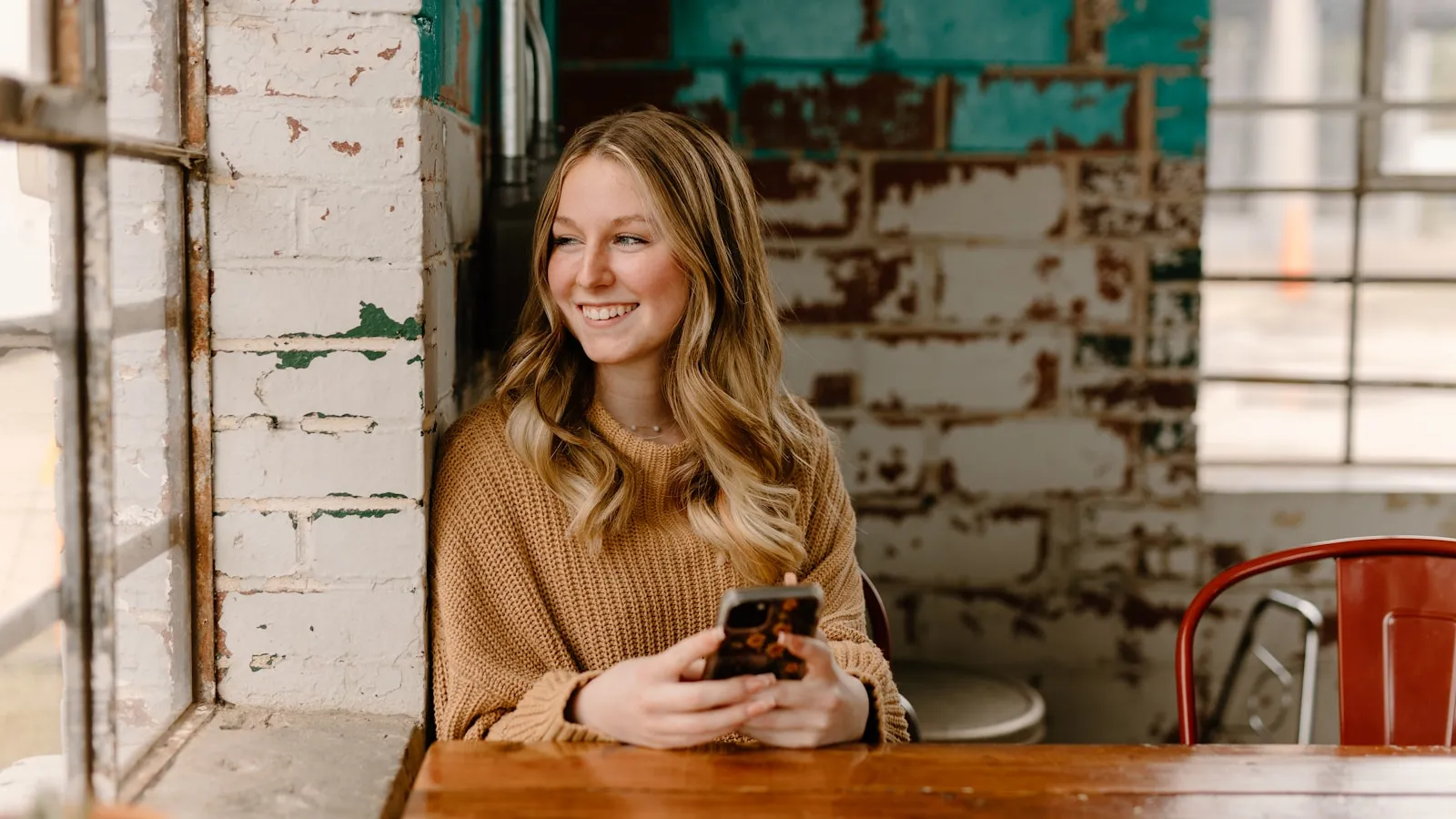 Vrouw met blond haar zit aan een tafel bij het raam met haar telefoon in haar handen.