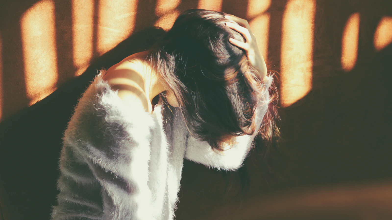 Vrouw met gebogen hoofd en een hand in haar haar bij warm invallend licht.