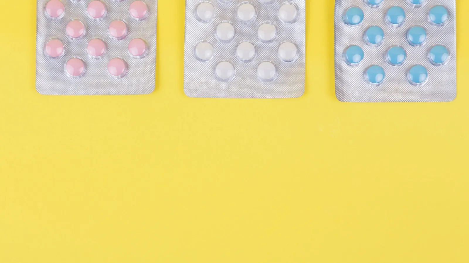 Drie doordrukstrips met roze, witte en blauwe tabletten op een gele achtergrond.