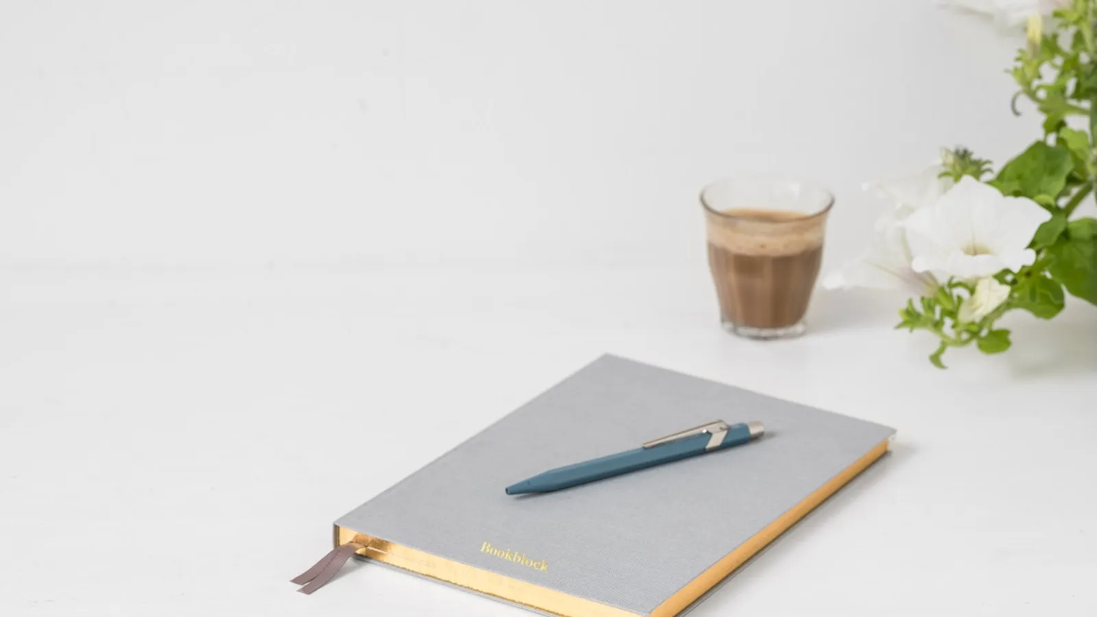Grijs notitieboek met een blauwe pen op een witte tafel, naast een glas koffie.