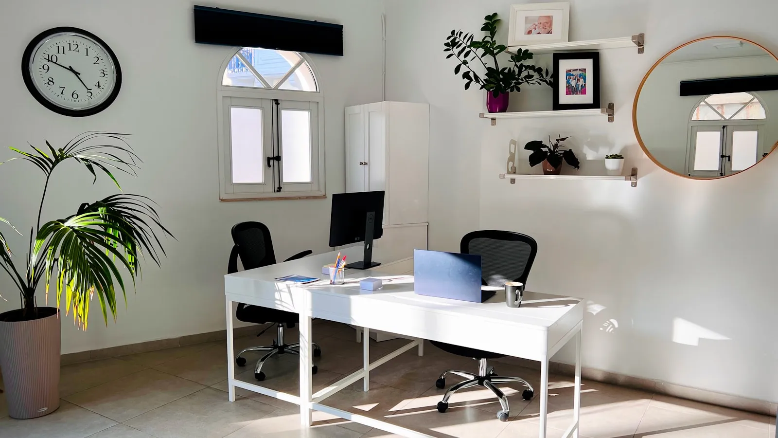 Lichte werkkamer met een ronde wandklok boven een witte bureautafel.