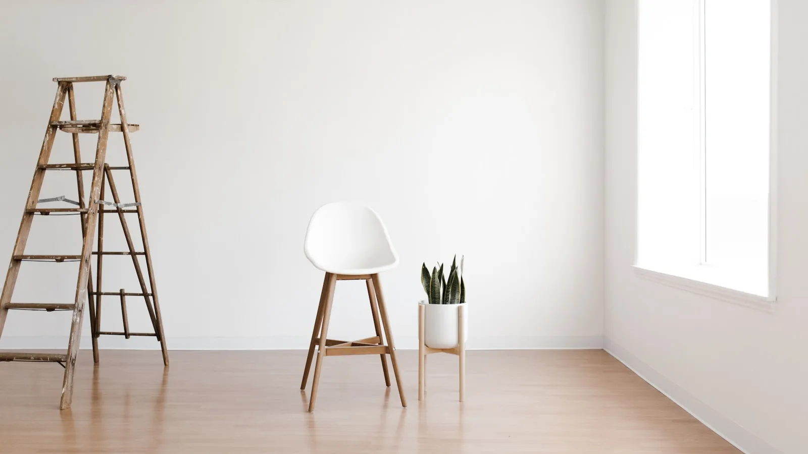 Lichte, lege kamer met een witte stoel, een houten trapladder en een plant bij het raam.