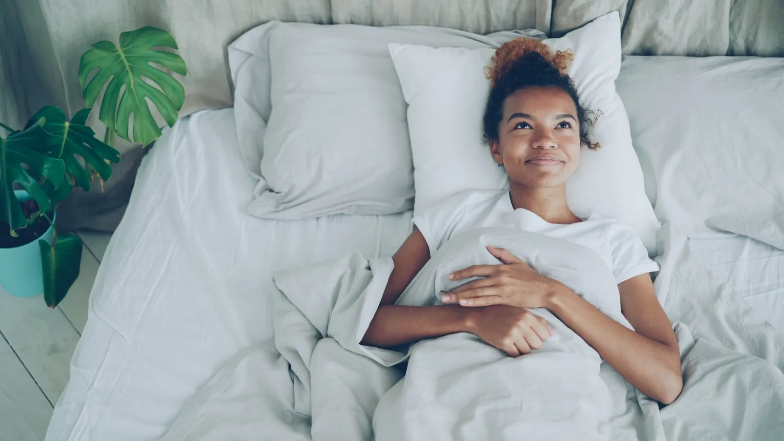 Een vrouw ligt wakker in bed met wit beddengoed, naast een kamerplant.