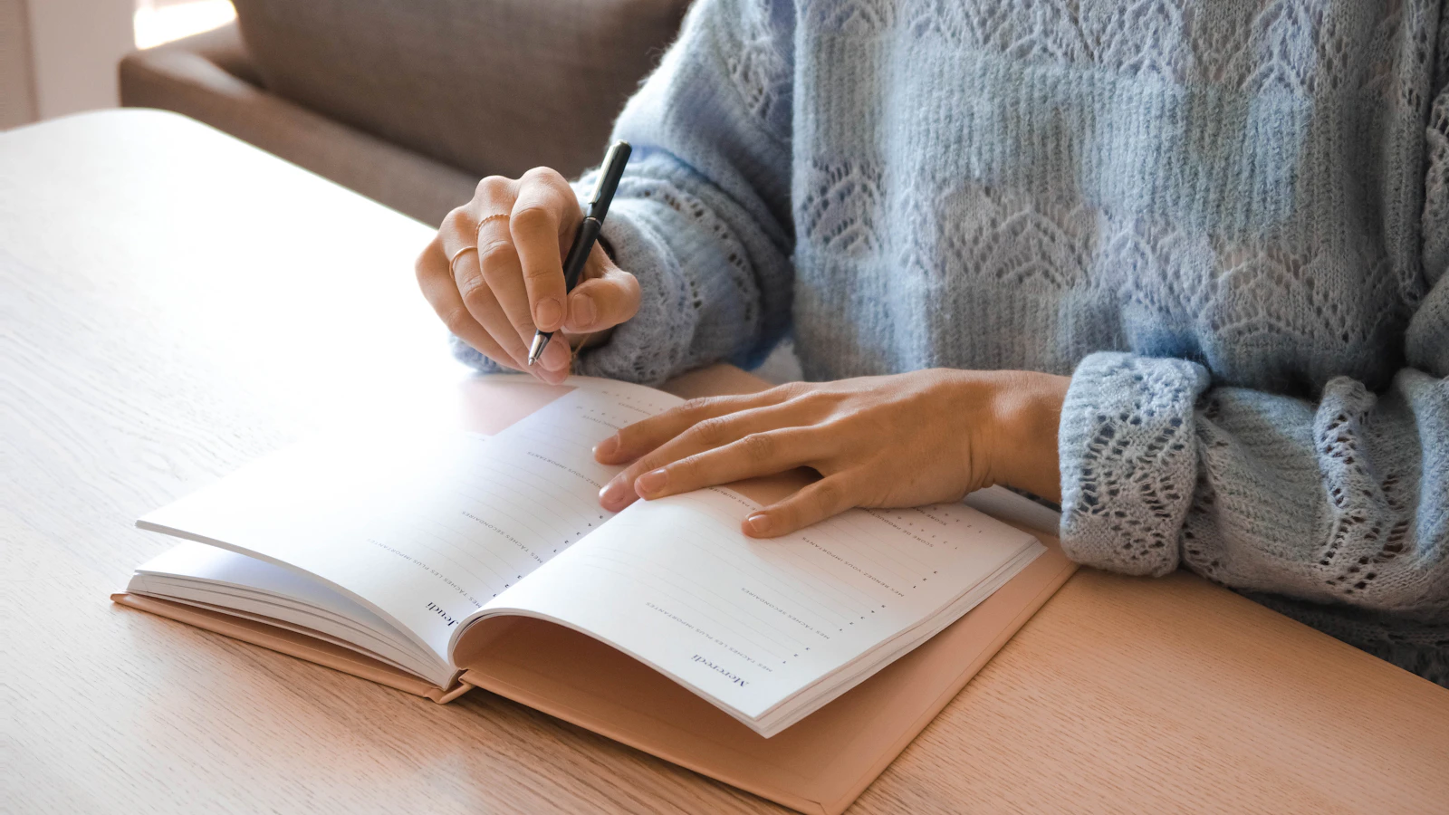 Vrouw in een lichtblauwe trui schrijft in een notitieboek aan tafel