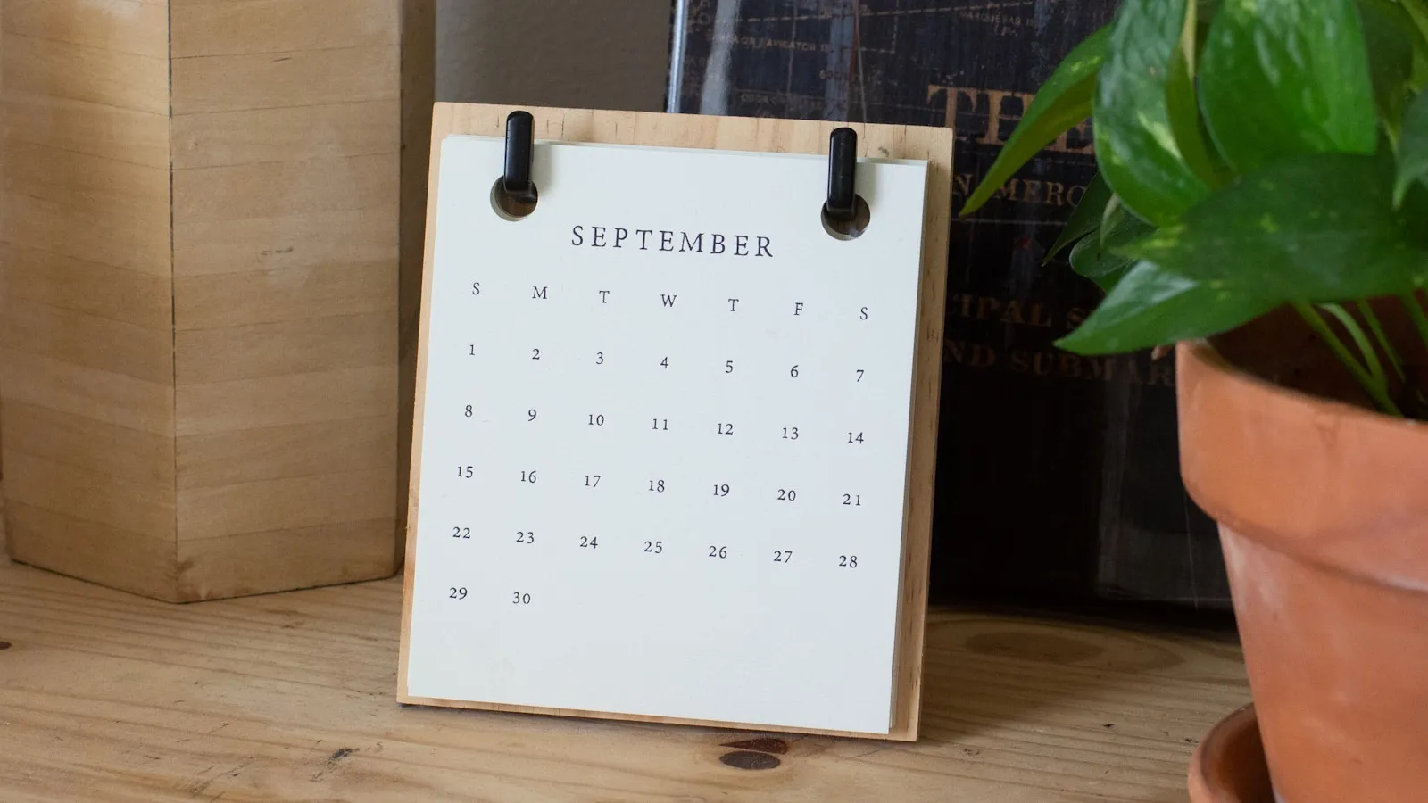 Papieren maandkalender op een houten tafel naast een plant.