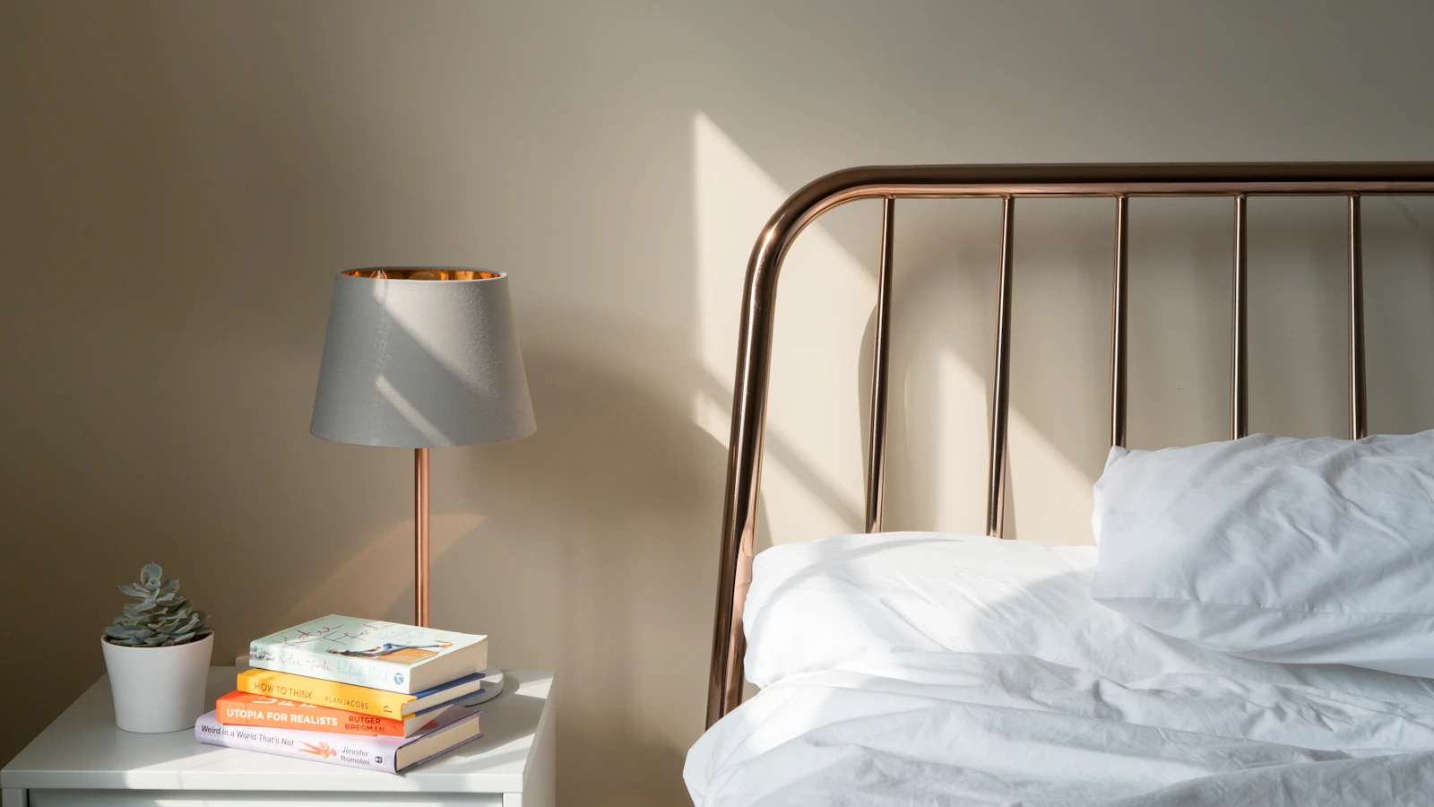 Nachtkastje met een lamp, een stapeltje boeken en een plantje naast een opgemaakt bed in het ochtendlicht.